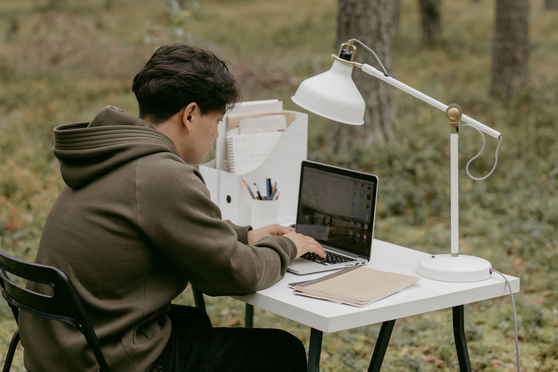 Man in hoodie using laptop at an outdoor workspace in a serene forest setting.