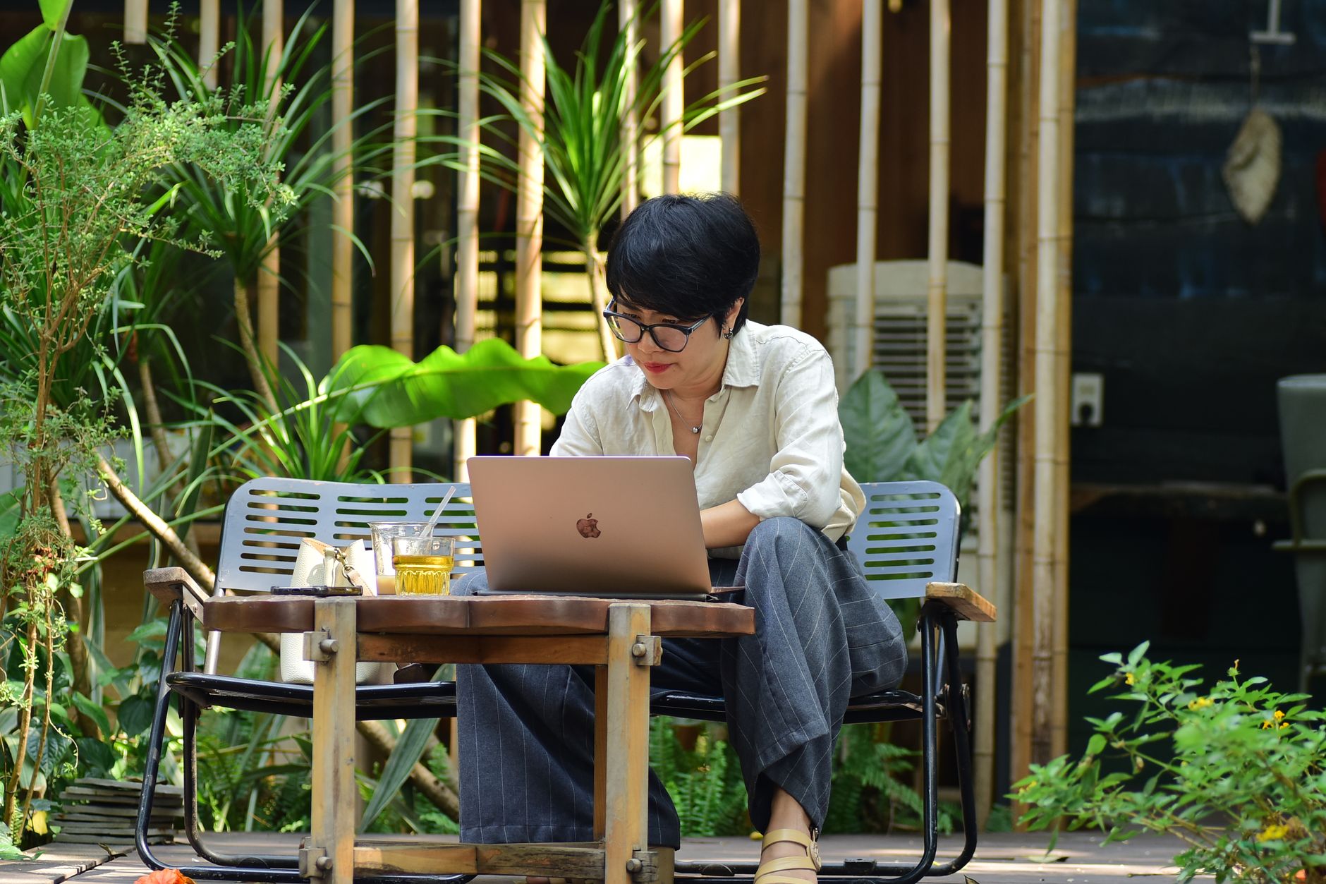 A woman focuses on her laptop while sitting in a lush outdoor cafe.