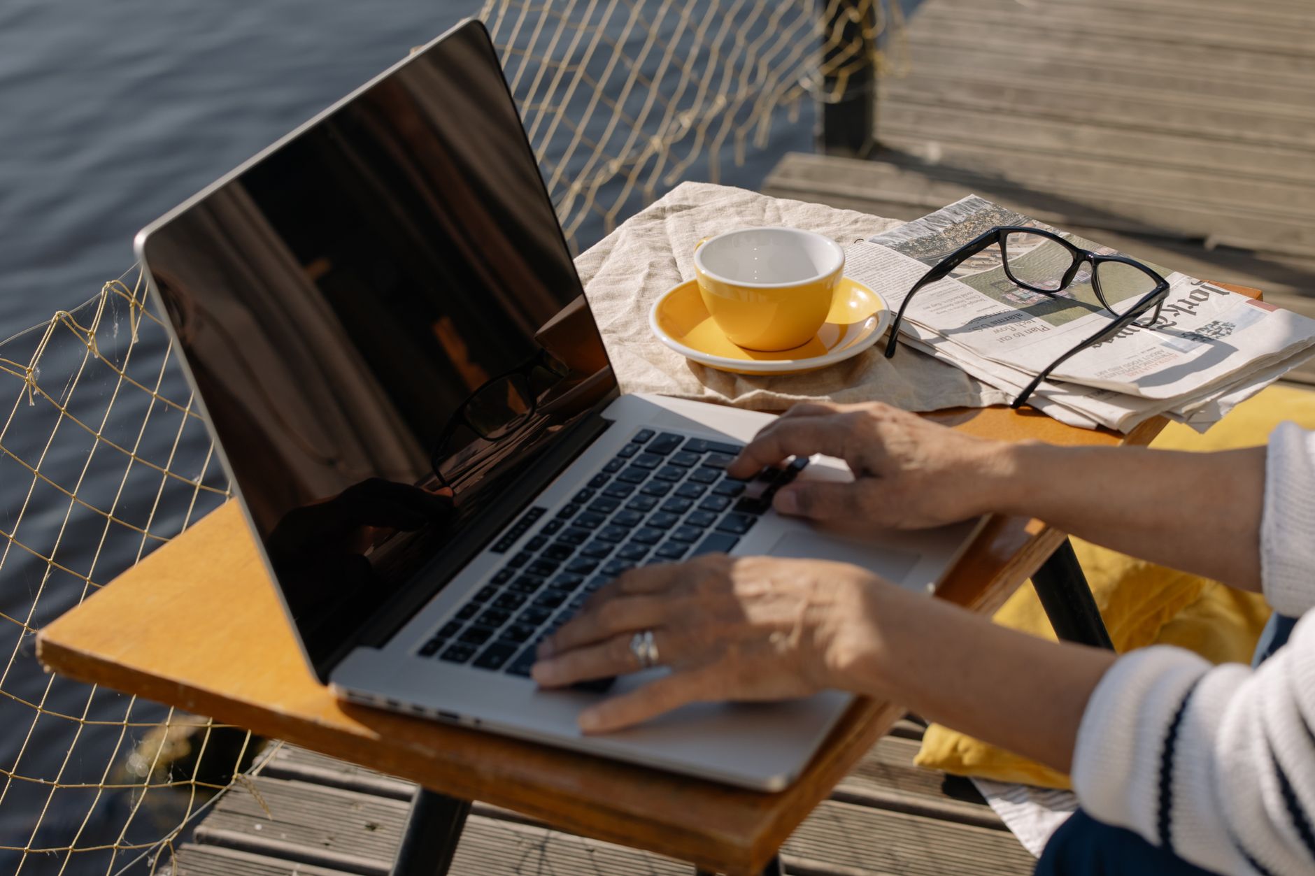 A person typing on a laptop outdoors by a lakeside with coffee and newspaper, ideal for remote work imagery.