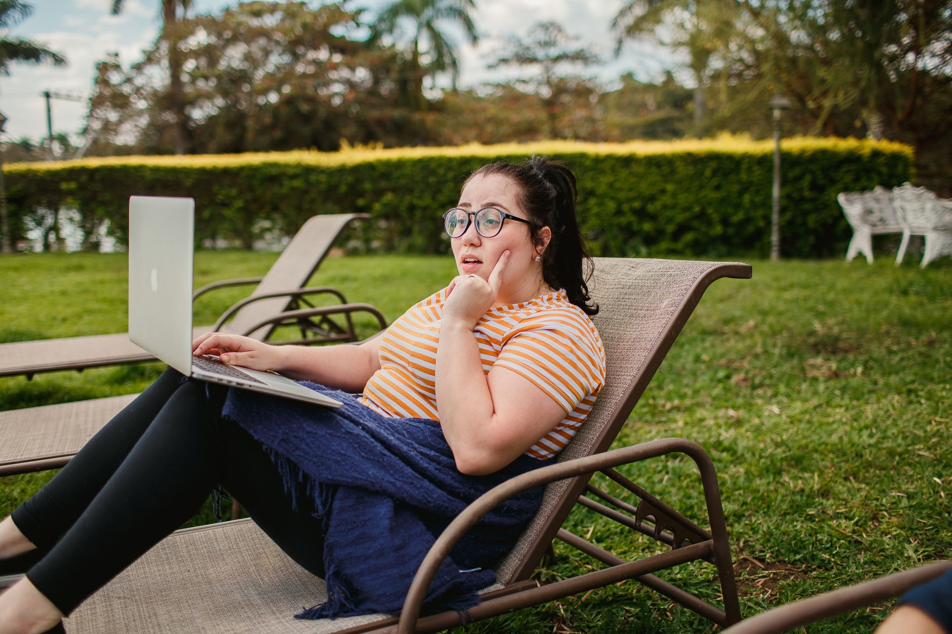 Woman relaxing on lounge chair with a laptop in a lush green garden, enjoying a sunny day.